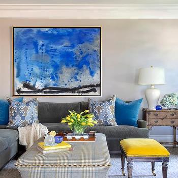 Blue living room with mustard yellow accent ottoman. There is a large blue and yellow abstract painting on the wall and a plaid, nail head ottoman.