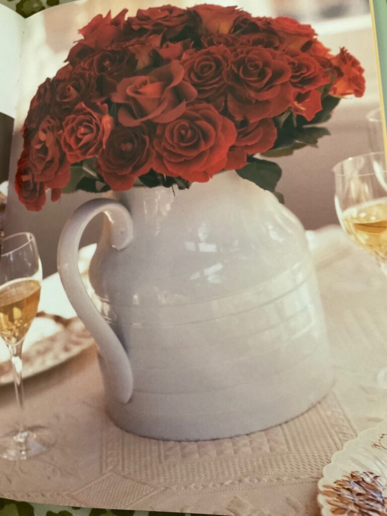A high-quality photograph of a white ceramic pitcher vase filled with deep red roses, positioned on a white textured tablecloth.