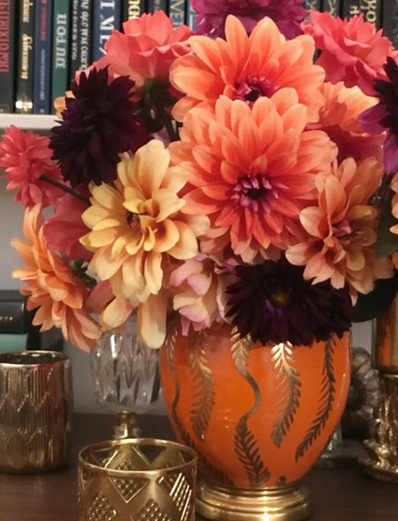 A close-up photograph of a floral arrangement in an ornate orange ceramic vase with black leaf designs. The vase contains a vibrant bouquet of coral pink dahlias, deep burgundy dark dahlias, and cream-colored chrysanthemums.