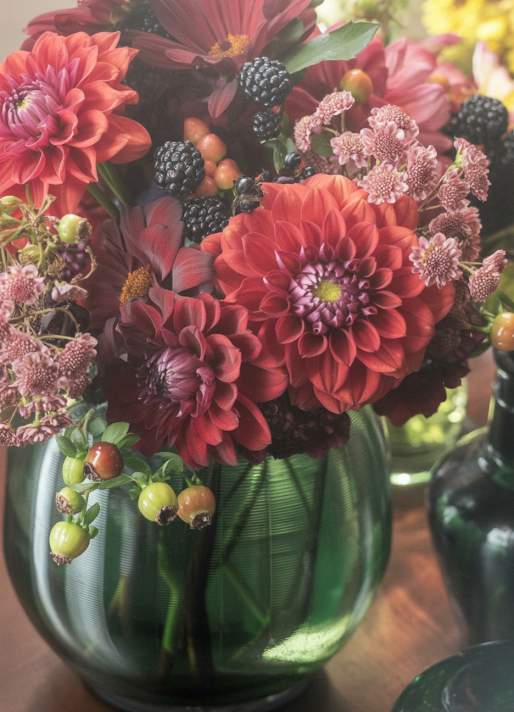 A close-up photograph of a vibrant floral arrangement in a dark green glass vase. The arrangement features large red dahlias with layered petals, deep burgundy chrysanthemums, and small pink button mum flowers. Fresh blackberries and small green berries are scattered throughout the arrangement.